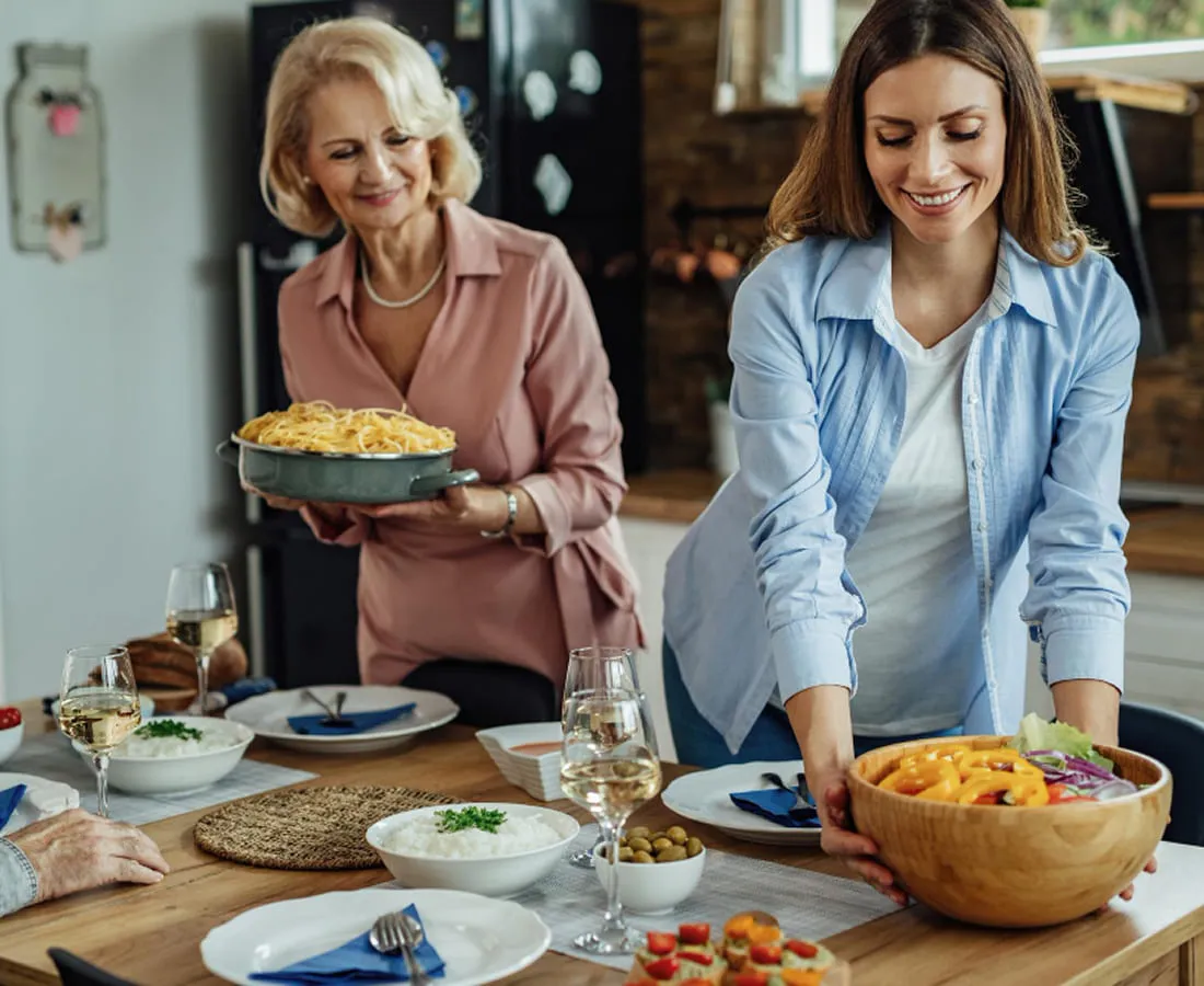 Two generations of a family bringing dishes to a laid dinner table