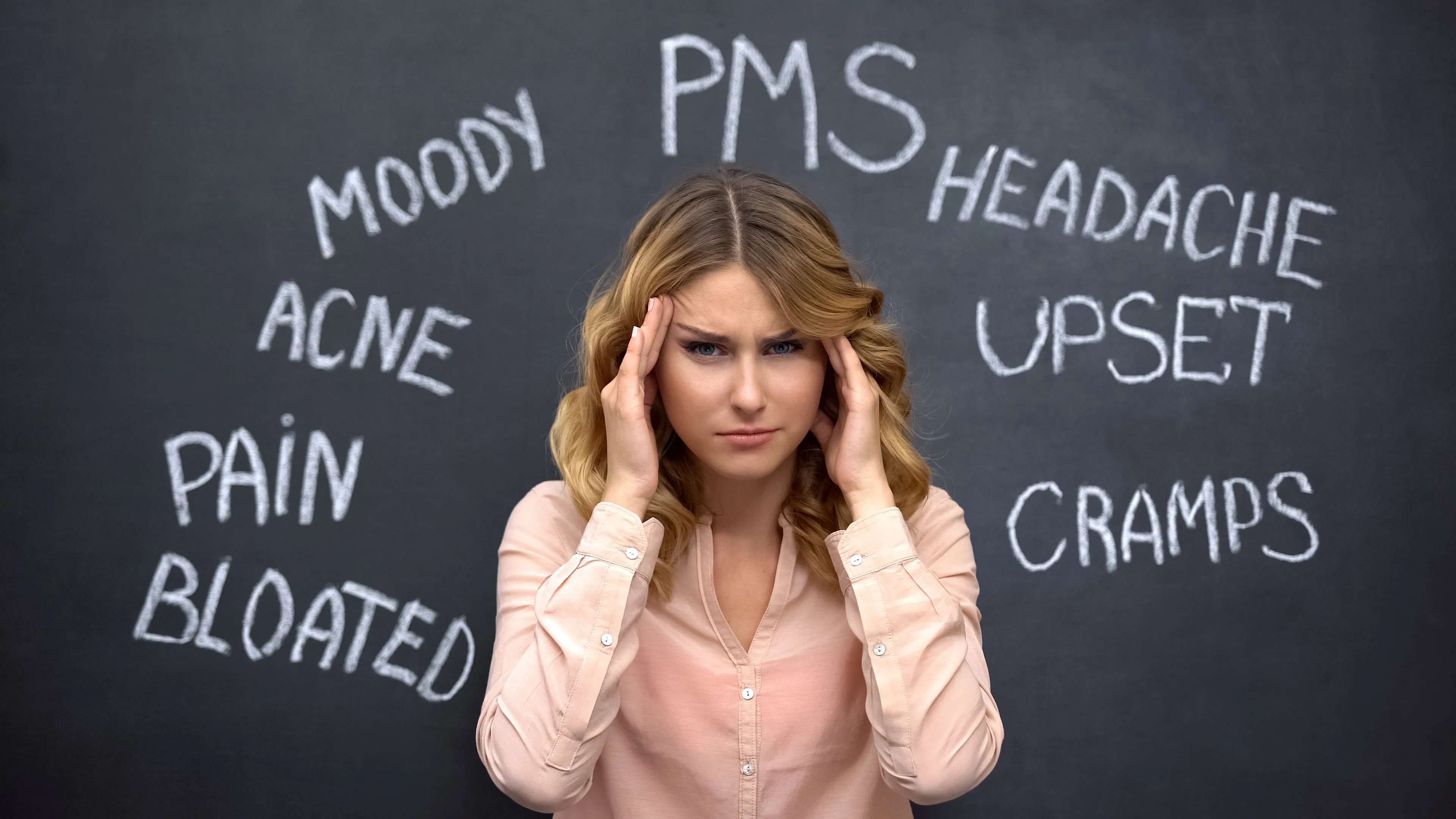 A woman holding her head in front of a blackboard chalked with PCOS symptoms: moody, headache, acne, cramps, bloated
